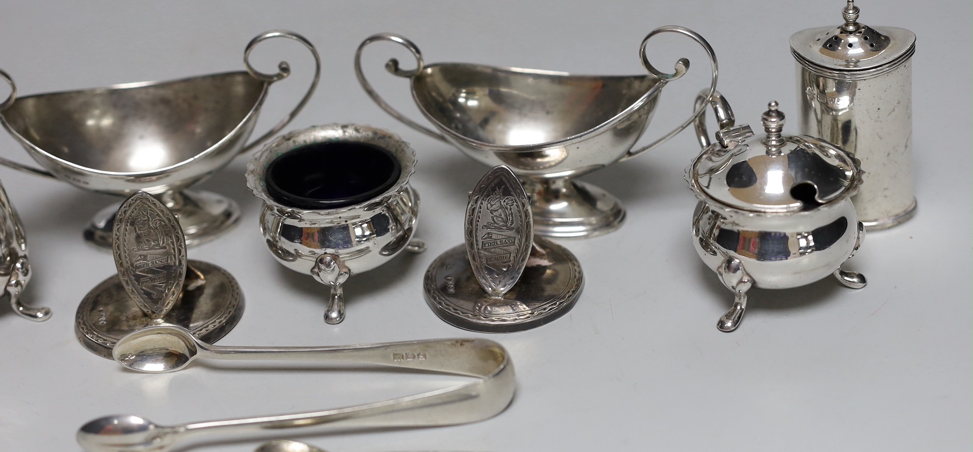 A pair of George V silver two handled boat shaped pedestal salts length 10.7cm, a pair of Edwardian silver pepperettes, a pair of late Victorian silver menu holders, a pair of silver sugar tongs and a three piece plated